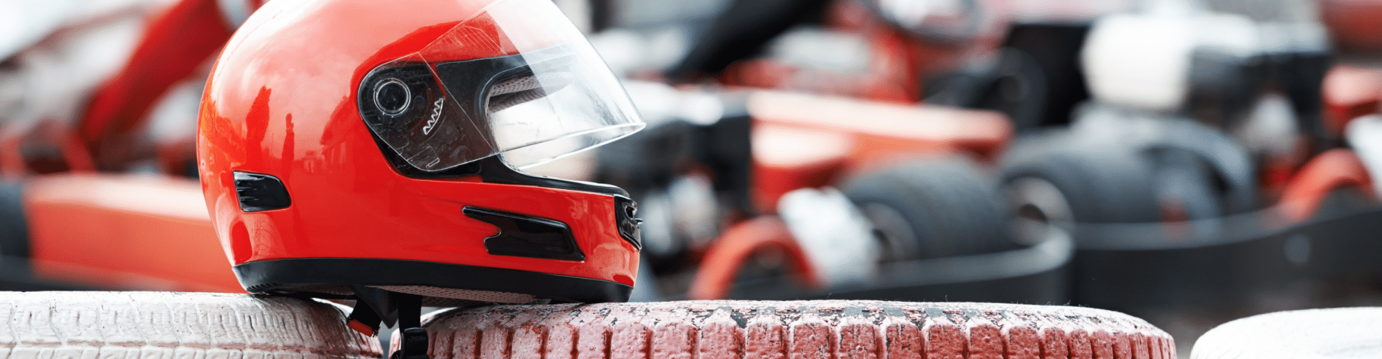 Roter Motorsporthelm liegt auf gestapelten, rot-weiß lackierten Reifen am Streckenrand. Im Hintergrund verschwommen mehrere Karts.