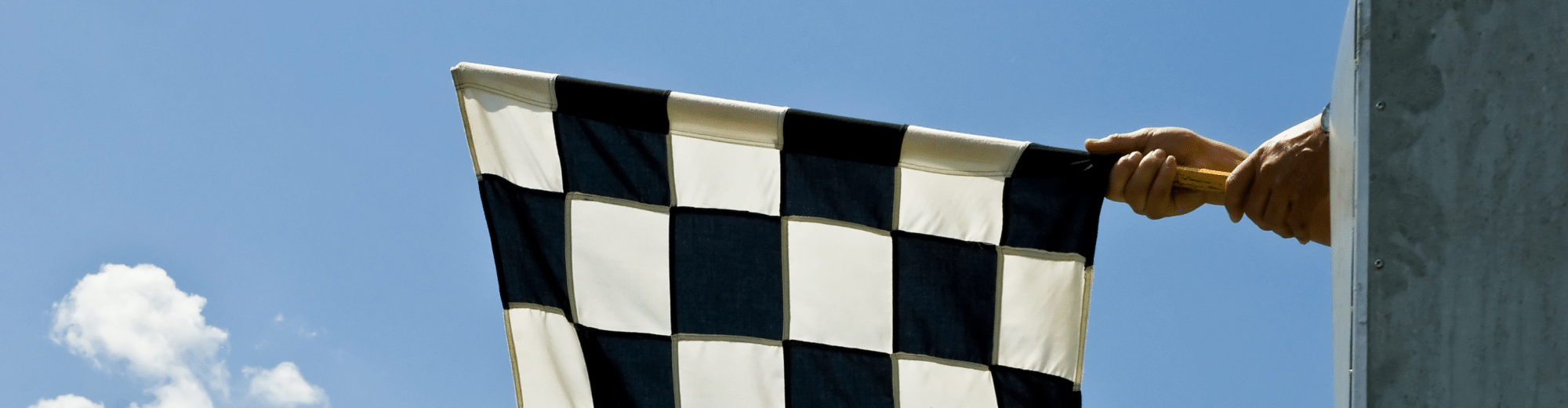 Eine schwarz-weiß karierte Zielflagge wird von einer Hand aus einem Podest heraus vor blauem Himmel geschwenkt – Symbol für das Rennende.